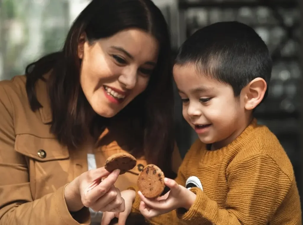 A smiling woman and young boy share a joyful moment holding chocolate chip cookies, suggesting warmth, comfort, and family connection, ideal for promoting autism-friendly vacations that prioritize sensory-friendly environments and positive shared experiences.